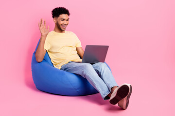 Full length photo of cheerful friendly guy wear yellow shirt sit on pouf waving palm on laptop...