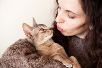 Close up of woman hugging and kissing her grey cat on a beige background. Cute Abyssinian kitten of blue color. Love relationship between human and cat. Cat day. Copy space. Selective focus.