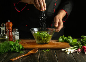 Professional chef salts chopped lettuce leaves in a plate. Working environment in a kitchen. Delicious salad with fresh vegetables