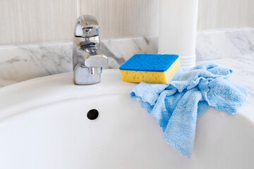 Cleaning the bathroom. A washing sponge, blue rag and a cleaning gel stand on a white sink in...