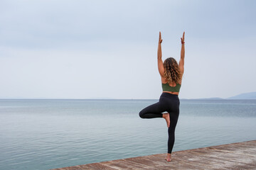 Woman practicing yoga by the sea on a deck - Vrksasana Urdhva Hastasana -  Tree pose hands up