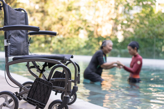 Selective Focus Off Wheelchair Background Of Physical Therapist Helping Senior Woman In Aqua, Old Women And Mature Doing Aqua Aerobics Exercise In Swimming Pool, Elderly Sports, And Active Lifestyle.