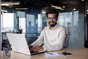 Portrait of a young Indian businessman working in the office at a laptop, smiling and looking at...