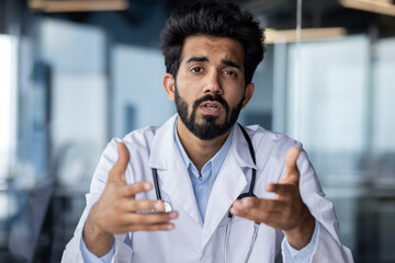 Online medical consultation. Portrait of a serious young Indian male doctor sitting in the office...