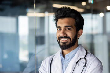 Close-up photo. Portrait of a young Indian male medical student, intern. Standing smiling and looking at the camera in a white coat and with a stethoscope