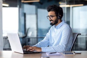 A young Indian businessman is working in the office on a laptop wearing a headset