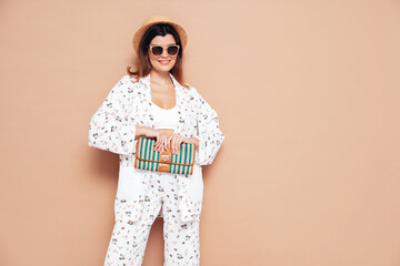 Young beautiful smiling female in trendy summer costume clothes. Carefree woman posing near yellow wall in studio. Positive model having fun indoors. Cheerful and happy. In hat. Holds clutch