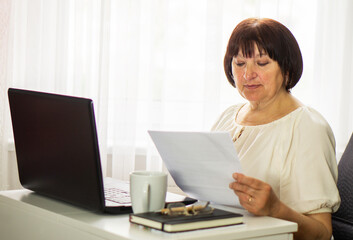Middle aged senior woman sitting with laptop and paper document, working and drinking coffee.