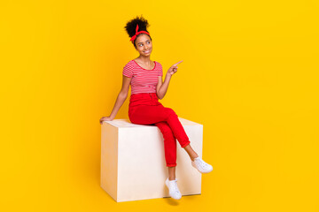 Full body photo of little schoolgirl sitting podium cube look direct finger empty space isolated on yellow color background