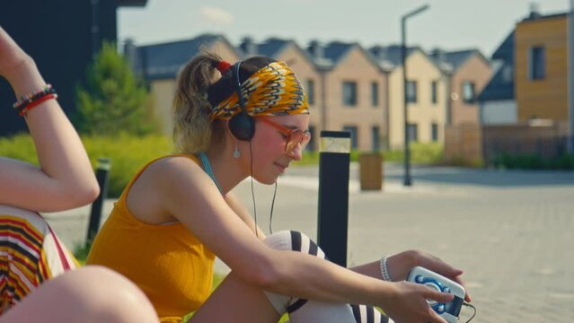 Medium shot of young Caucasian girl in bright hippie style clothes wearing headphones and listening to old CD player while sitting on green lawn with friends