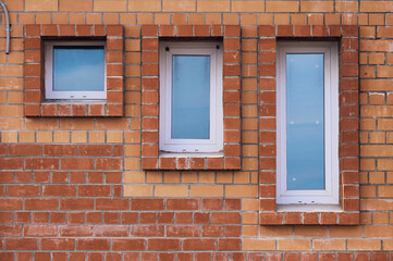 Minimalistic shot of a brick wall with windows of various sizes. Three windows with plastic frame. Red brick masonry. Facade of a building of unusual design.