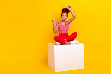Full size photo of funky young girl sit platform point empty space dressed stylish red striped clothes isolated on yellow color background