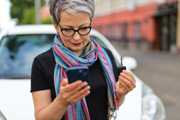 A senior woman with a smartphone in her hands and car keys uses a rental app. - Powered by Adobe