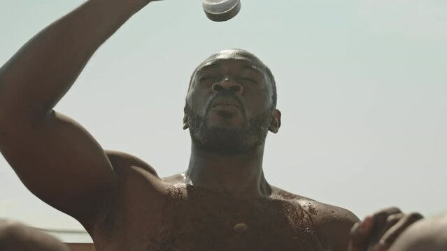 Low angle shot of muscular shirtless black man sitting outdoors on sunny day, drinking and pouring water from bottle over his head while resting after intensive physical training