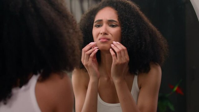 Worried young beautiful African American woman sad ethnic girl looking in mirror stressed about facial wrinkles problem acne frustrated upset touching face dry sensitive skin care facial blackheads