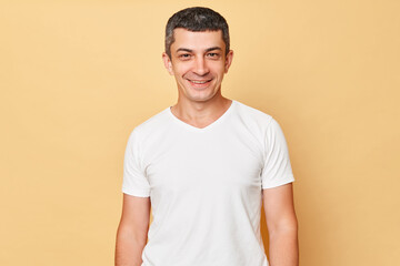 Fototapeta premium Smiling handsome man wearing white casual t-shirt standing isolated over beige background looking at camera being in good mood expressing positive emotions.