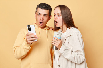 Surprised beautiful woman and handsome man wearing casual style clothing standing isolated over beige background browsing internet together on smartphone drinking hot coffee from thermos.