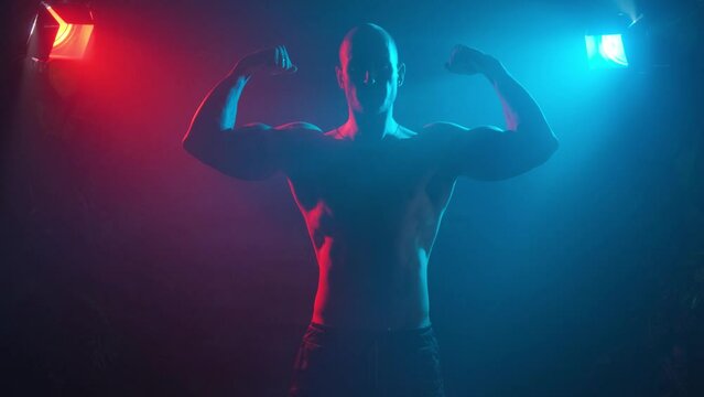 Male bodybuilder shows muscles on his arms in neon light in smoke.