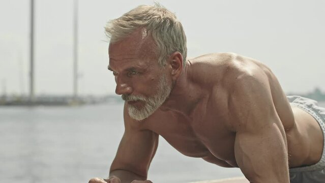 Tilt up shot of senior gray-haired man with muscular shirtless torso holding forearm plank while training outdoors on summer day
