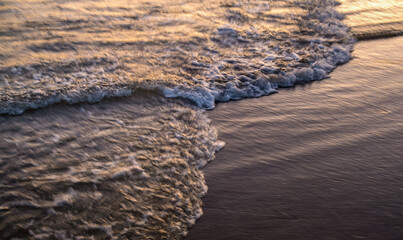The oncoming wave in the blur in the sunset light. Background of water trail in long exposure with white foamy splashes.