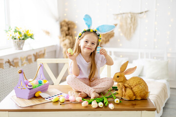 easter, cute little girl child with bunny ears on her head playing with colorful easter eggs on a table in a bright room, lifestyle,