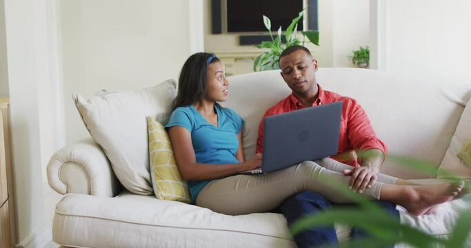 Happy biracial couple sitting on sofa with laptop and talking
