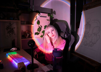 A blonde girl in her room at the computer is sitting in a chair in a rainbow light