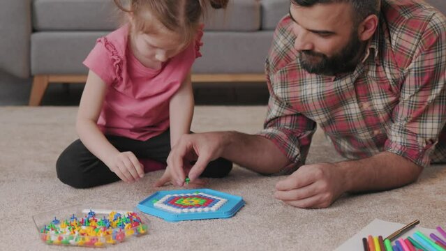 Young handsome father and cute daughter playing together. Dad and child making a puzzle with mosaic pegboard lying on the carpet at home. Educational toys for children. Home leisure. Father's day.