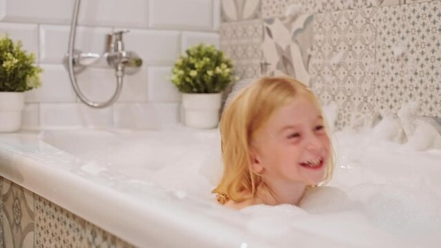 Laughing caucasian blonde three years old girl taking bath with white foam bubbles, playing, having fun, looking at camera.Carefree childhood, body washing, hygiene concept.