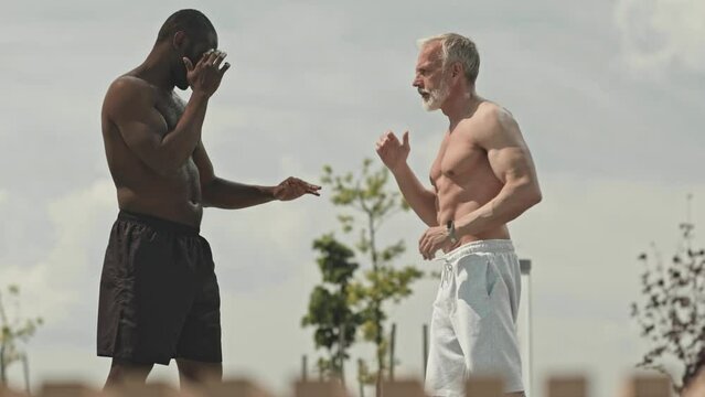 Medium long shot of muscular African American trainer teaching senior Caucasian man kickboxing technique during outdoor class on street