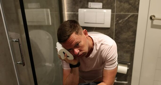 Man applies toilet paper to head sitting on toilet bowl near shower. Concept of daily routine in light bathroom of accommodation slow motion