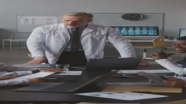 Vertical shot of mature Caucasian male head physician having conference meeting with his multiracial medical team sitting with paper documents and laptops at desk in modern office