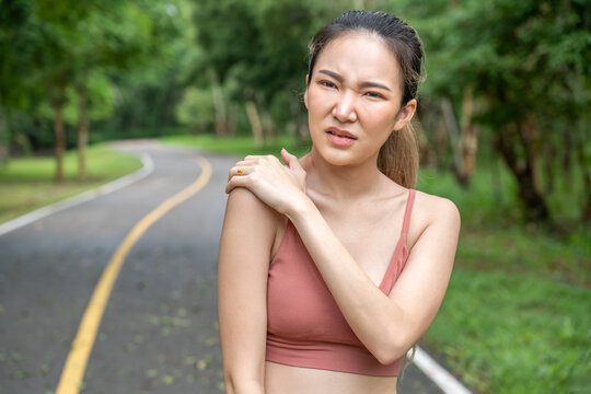 Young atractive Asian woman in fitness clothes putting her left hand over her pain right shoulder while standing at running track of a local park