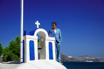 Elegant handsome groom in blue suit is posing near the Church in Santorini, Oia. Engagement and...