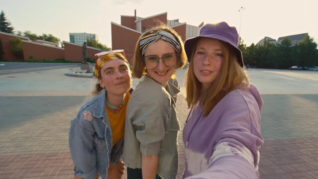 Medium shot of three cheerful bright teenage girls looking at camera, smiling, posing and taking selfie outside in street at daytime