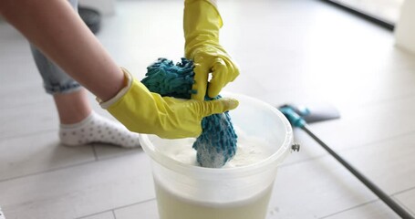 Woman in rubber gloves wrings foamy mop rag over bucket with water in room. Person works with floor house cleaning supplies slow motion