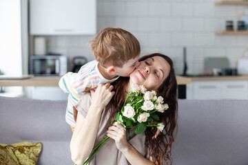 Little boy hugging and kissing his mother and giving her a bouquet of roses at home. Mother's day concept. Selective focus.