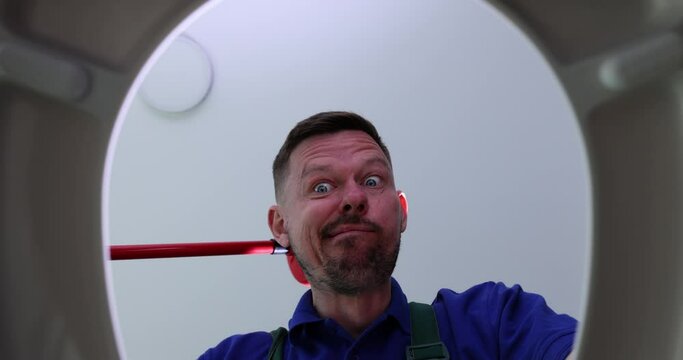 Funny plumber opens toilet bowl and scratches nape with plunger in restroom. Emotional man cleans clog in pipe with traditional tool view from bottom