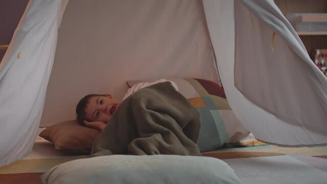 Cute little boy with down syndrome lying under soft blanket on pillow in cozy indoor playhouse