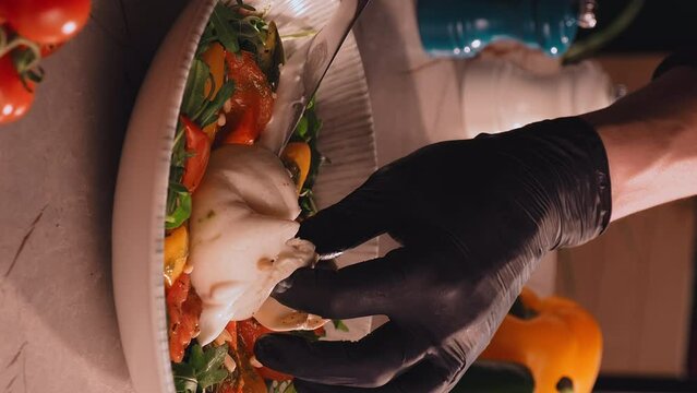 Closeup shot of burrata salad. Chef cuts burrata cheese served with tomatoes, yellow pepper, aragula and pine nuts in a plate. Italian appetizer. Vegetarian lifestyle concept