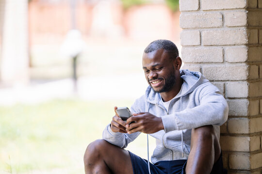 Cheerful black man sitting on floor of building and browsing smartphone