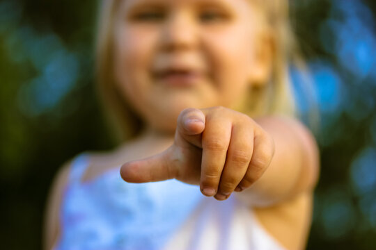 A little blonde infant girl pointing finger into a distance straight ahead. Baby girl's hand in focus. Age 2-3 years old. Cute children in a green park blurry background at summer day. Smiling kid.