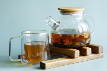 Glass mug and teapot with hot black flavored tea with fruits and flower petals on a wooden hot plate. Green-blue background. Copy space. Selective focus