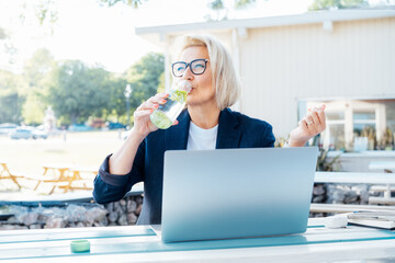 Healthy habit to drink water. Middle aged business woman drinking water with lemon and mint from the bottle during working remotly in park. Control body hydration, Tracker water balance. - Powered by Adobe