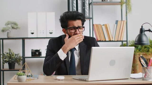 Bored sleepy indian businessman worker working on laptop computer, yawning, leaning on hand falling asleep at office. Exhausted tired freelancer workaholic young man. Employment, occupation, workless