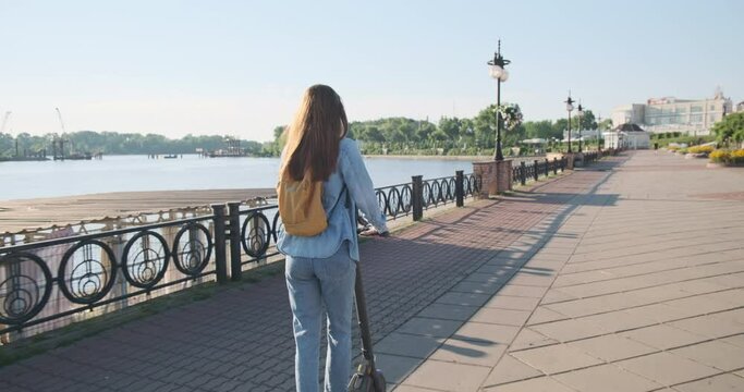 Red-haired young woman kick start rides electric scooter in park, river embankment, tracking camera, back view.