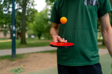 An elderly man next to a blue ping pong table hits an orange ball on a tennis racket in a city park