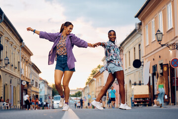 Carefree female friends hold hands while dancing on street.