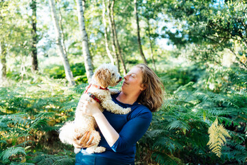 Happy middle-aged woman embracing her cockapoo puppy pet dog during walk outdoors in summer forest or park on sunset. Cute little doggy. A woman petting her dog like a small child. Selective focus