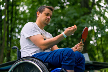 Inclusiveness A disabled man in a wheelchair with a racquet and a ping-pong ball in his hands near a tennis table in a park 